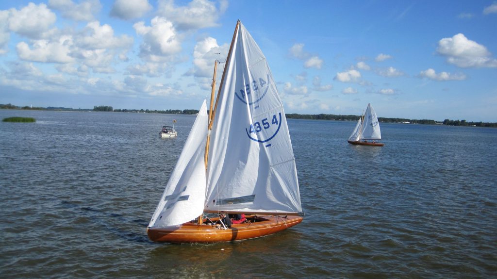 Houten zeilboot met witte zeilen op blauw water onder een wolkenlucht