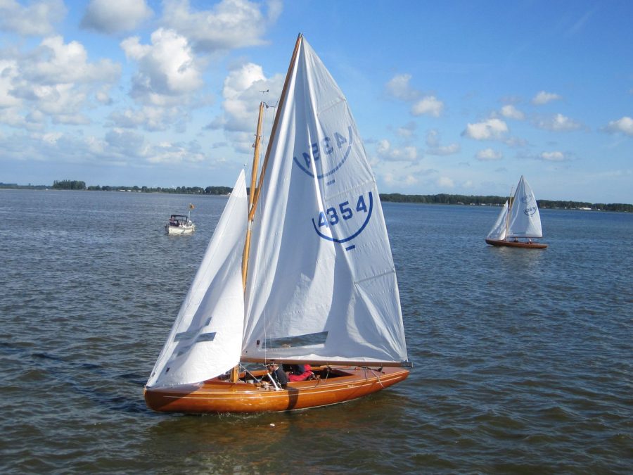 Houten zeilboot met witte zeilen op blauw water onder een wolkenlucht